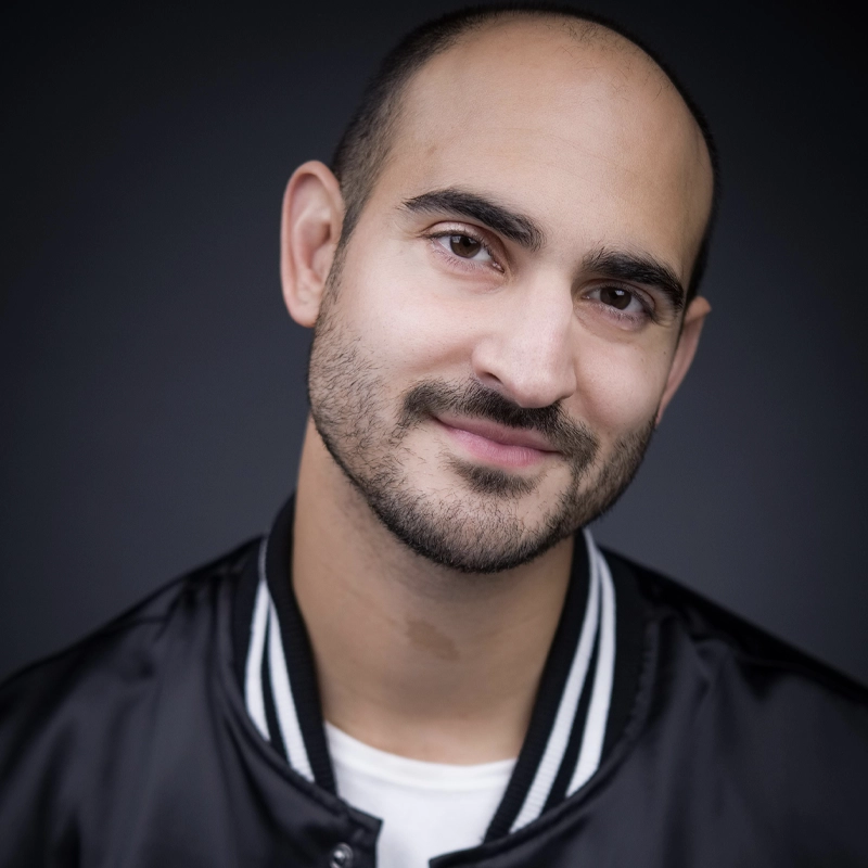 A close-up portrait of performer Nathan Lucrezio with a shaved head and dark stubble, smiling warmly at the camera. He wears a black bomber jacket with white striped trim over a white shirt against a dark grey background.