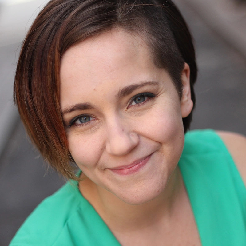 A young woman with light skin and short brown hair, partially shaved on one side, smiles warmly at the camera. She wears a bright green top and has blue eyes. The softly blurred background hints at an outdoor setting, captured by Anthony Alfaro.