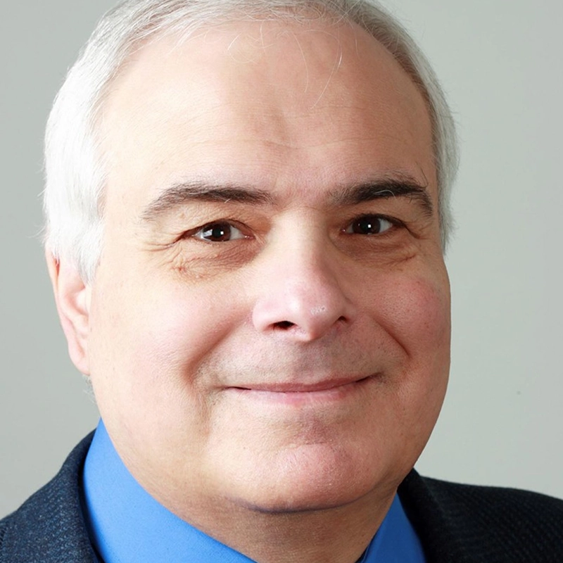A middle-aged man with short white hair and fair skin, Peter Filichia smiles slightly at the camera. He is wearing a blue dress shirt and a dark blazer. The background is plain and light gray.