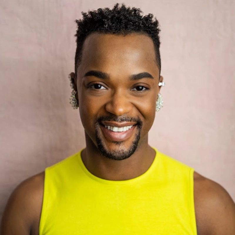 A smiling J. Harrison Ghee with short, curly black hair and neatly groomed facial hair wears a sleeveless bright yellow top and silver ear cuffs. They stand against a pale pink background, looking directly at the camera with a friendly expression.