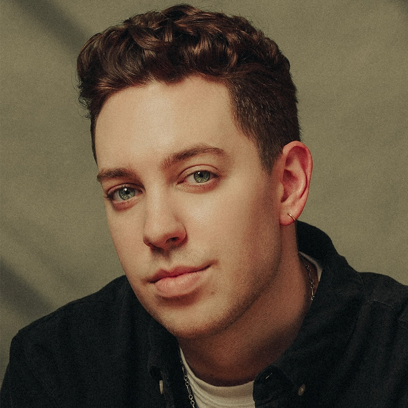 Jordan Jacobs, a young man with short, wavy brown hair and green eyes, looks at the camera with a slight smile. He wears a black collared shirt, a thin chain necklace, and a small hoop earring in his left ear against a softly lit background.