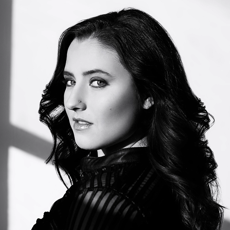 A woman, reminiscent of Kailey Marshall, with wavy dark hair and light eyes looks over her shoulder, wearing a striped top. The image is in black and white, with strong shadows cast on the wall behind her, highlighting her features and creating dramatic contrast.