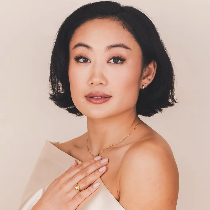 A woman with short black hair and light makeup poses against a beige background. Marina Kondo wears a strapless cream-colored top, a gold necklace, small earrings, and a ring, her hand gently on her chest and her expression calm and confident.
