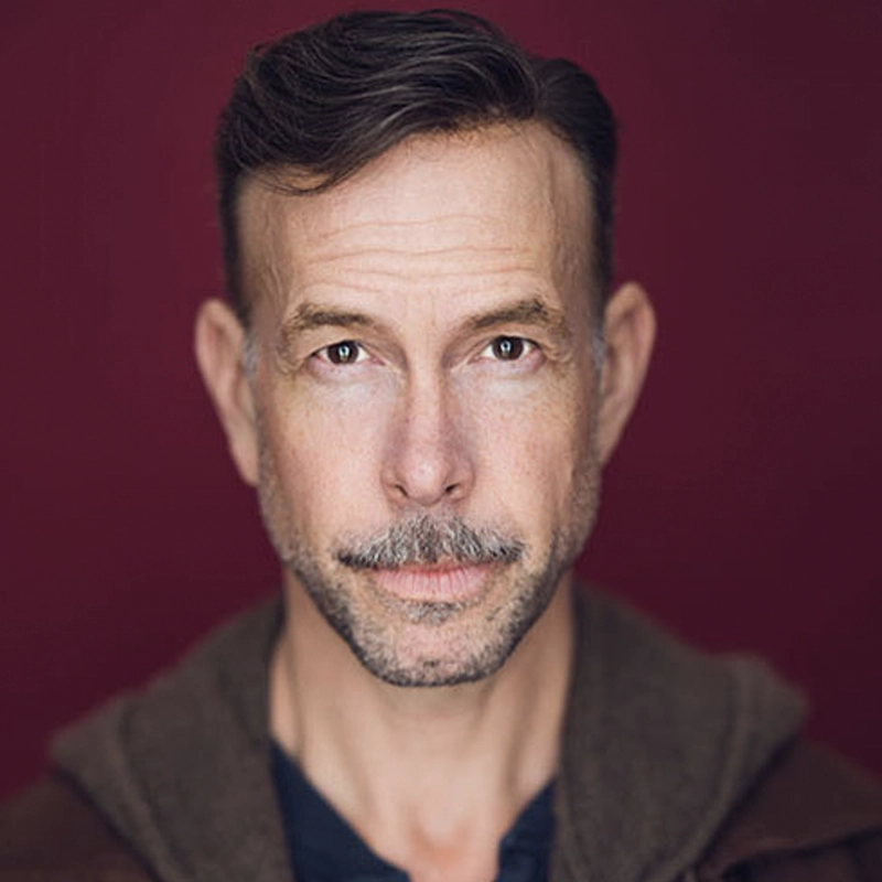 Wade McCollum, a middle-aged man with short brown hair, a trimmed mustache, and goatee, gazes directly at the camera. He wears a brown jacket over a dark shirt; the softly lit burgundy background highlights his facial features.
