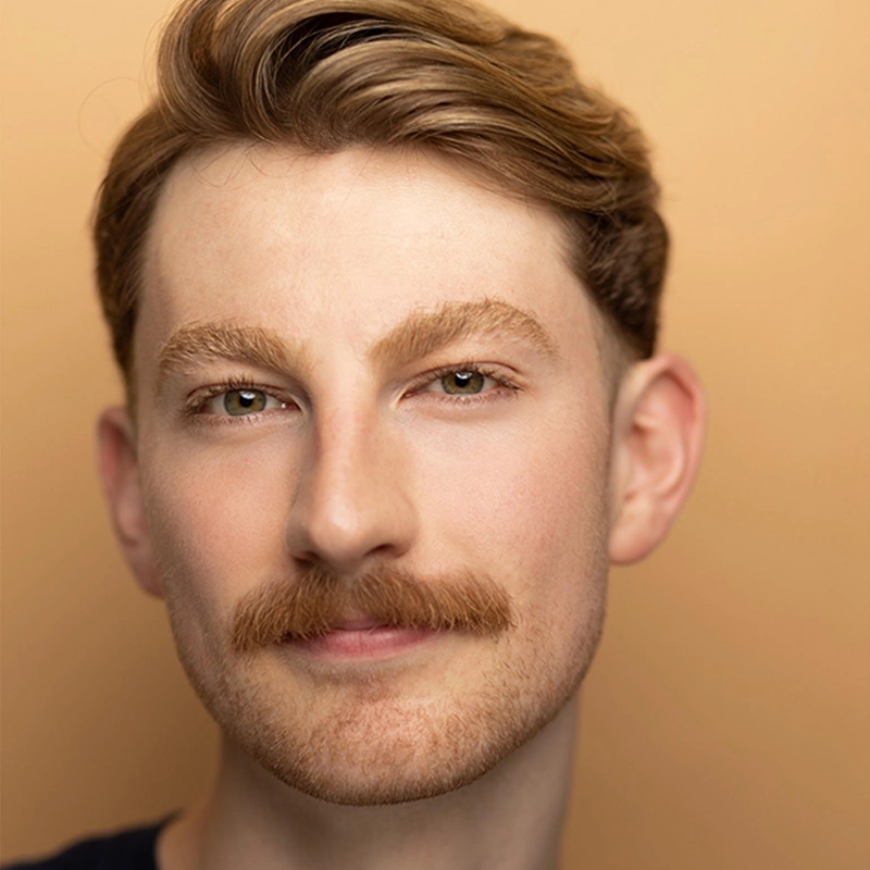 Jordan Jacobs, a young man with fair skin, light brown hair, and a thick mustache, smiles slightly. He has hazel eyes, neatly groomed eyebrows, and wears a dark shirt against a soft beige background that adds warmth to the portrait.