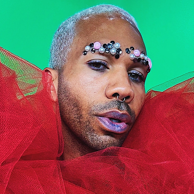 A person with short, curly gray hair poses against a bright green background. Reminiscent of Crystal Monee Hall’s bold style, they wear dramatic red tulle and have ornate gems and beads on their eyebrows, plus purple lipstick and subtle purple eyeshadow.