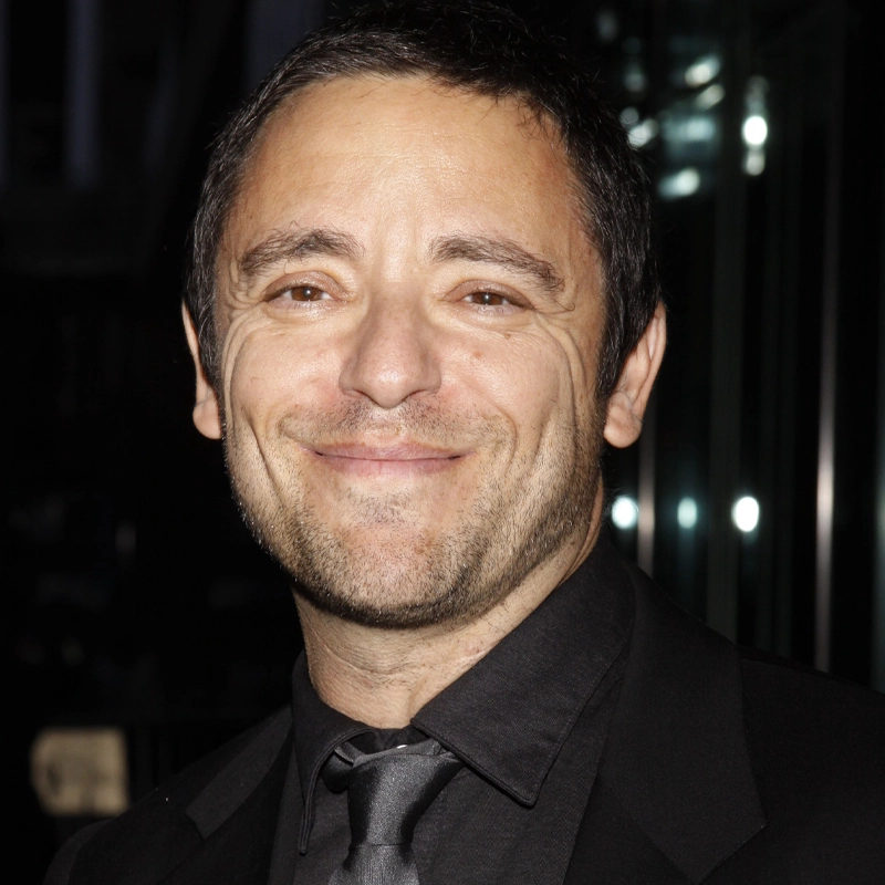 A man with short dark hair and a trimmed beard smiles softly. Wearing a black suit and metallic gray tie, he appears at an evening event—perhaps Steven Brinberg performing his acclaimed Simply Barbra, the ultimate Barbra Streisand tribute.