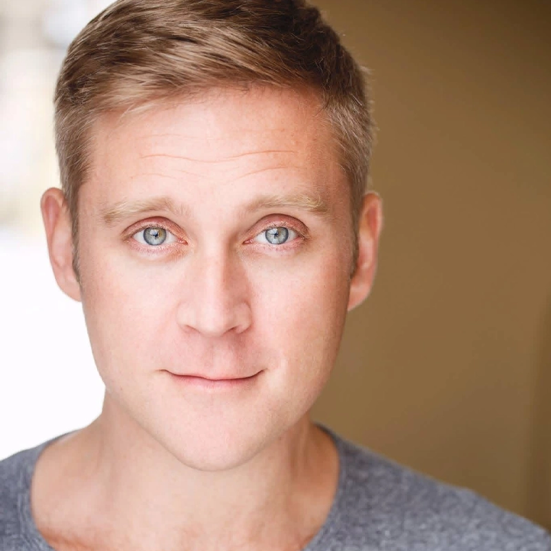 Michael Buchanan, a man with short light brown hair and blue eyes, looks directly at the camera with a slight smile. He is wearing a gray shirt, and the softly blurred warm-toned background creates a gentle and inviting atmosphere.