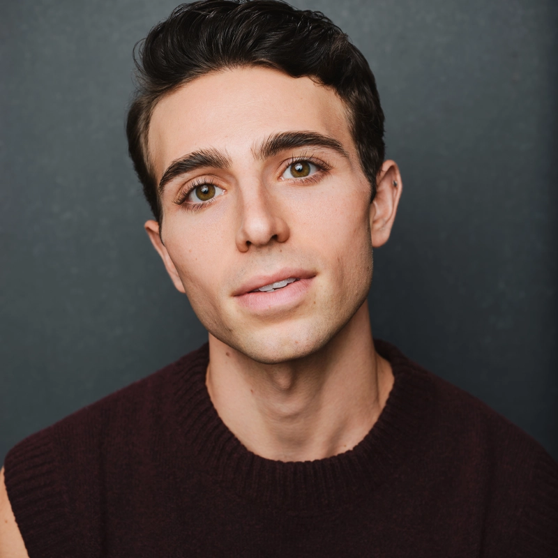 A young man with short dark hair, hazel eyes, and clear skin is wearing a sleeveless maroon sweater. Dean Cestari faces the camera with a slight smile, tilting his head gently, against a plain dark background.