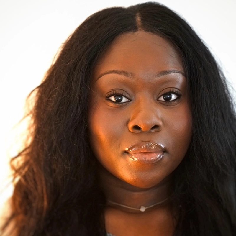 A woman with long, wavy black hair looks directly at the camera. She has smooth brown skin, glossy lips, and wears a delicate choker necklace. The softly lit white background highlights her face and hair in true Max Sangerman style.