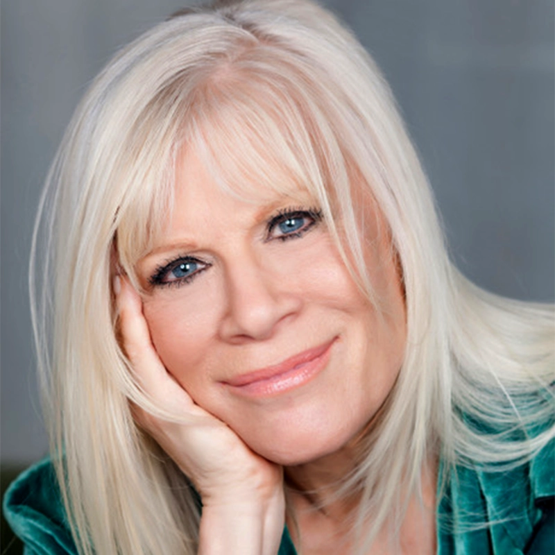 A smiling older woman, resembling Ilene Kristen, with straight, light blonde hair and blue eyes rests her chin on her hand. She is wearing a teal top, and her expression is warm and relaxed. The background is softly blurred.