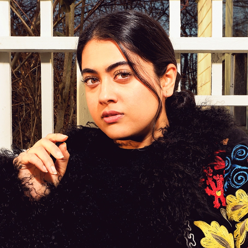 A young woman with dark hair parted in the middle poses outdoors in sunlight. She wears a black textured coat with colorful floral embroidery and looks directly at the camera. Pale fencing and bare tree branches appear in the background.