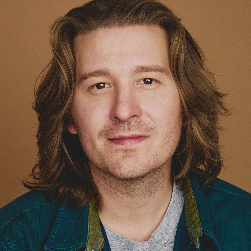 Max Sangerman, with wavy, shoulder-length light brown hair and fair skin, looks directly at the camera with a neutral expression. He wears a teal jacket over a gray shirt against a plain, warm brown background.