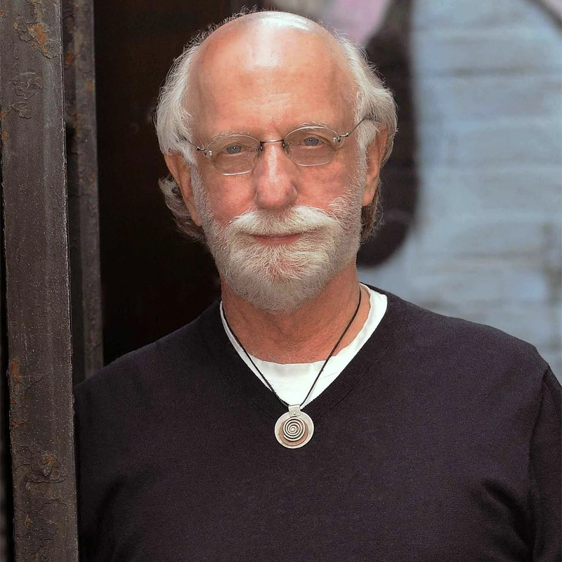 A portrait of a fair-skinned older man with a white beard and receding hairline, wearing wire-rimmed glasses and a black V-neck sweater. A silver spiral pendant on a black cord hangs around his neck against a white shirt. He stands against a backdrop of a textured, grey-blue wall.
