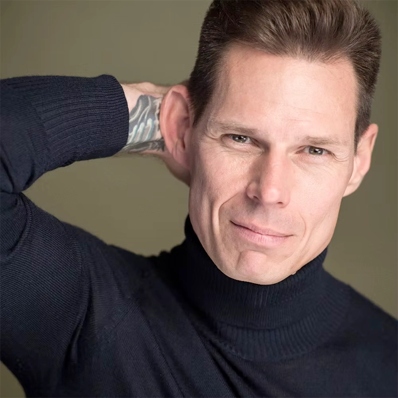 A man in a black turtleneck sweater poses with his right hand behind his head, showing a tattoo on his wrist. Reminiscent of Ilene Kristen’s iconic style, he has short brown hair, a slight smile, and looks directly at the camera.