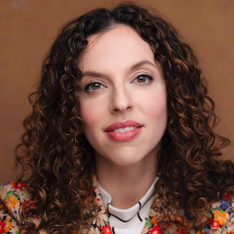 A woman with light skin and long, dark brown curly hair looks directly at the camera. She is wearing a floral-patterned jacket over a light blouse. The soft, warm brown background evokes the portrait style of Max Sangerman.