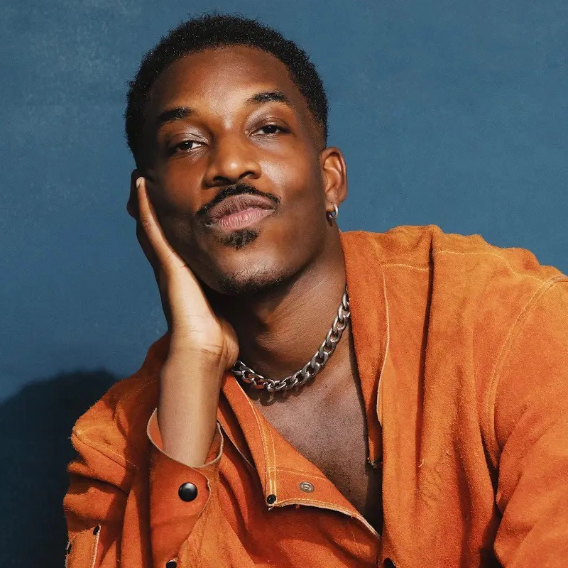 Britton Smith, a person with short hair and a mustache, rests their face on their hand, smiling slightly. They wear an orange suede shirt, a chunky silver chain necklace, and sit in front of a dark blue background. The shirt’s collar is open.