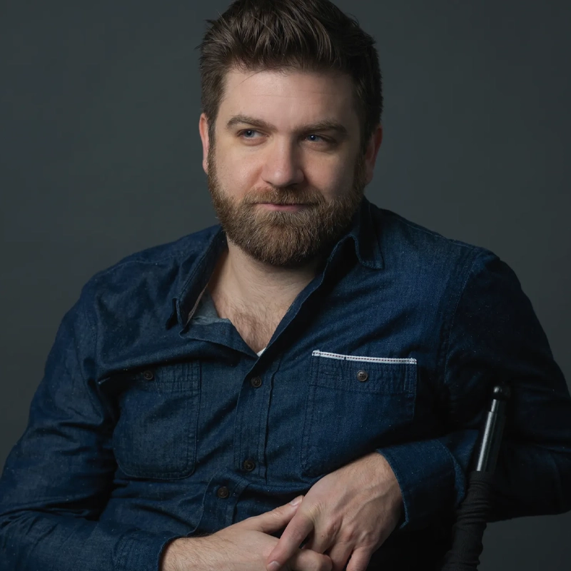 A man with short brown hair and a full beard, resembling Harris Doran, wears a dark denim button-up shirt as he sits against a dark gray background, gazing slightly to the left with a relaxed, thoughtful expression and his hands resting in his lap.
