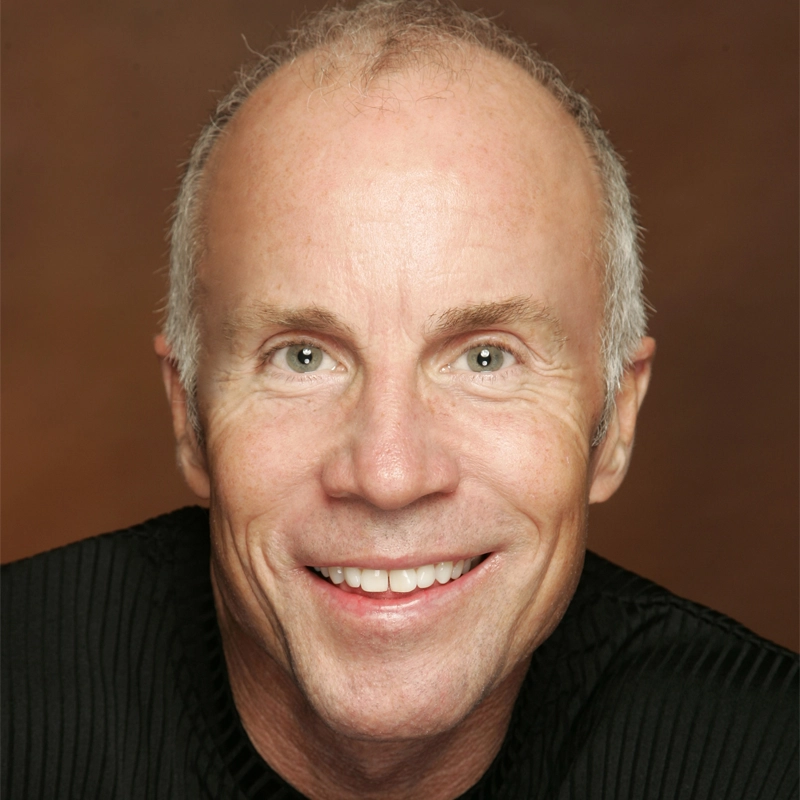 A smiling older man with blue eyes, light skin, and short, thinning gray hair is shown against a warm brown background. He is wearing a black, ribbed sweater and looking directly at the camera.