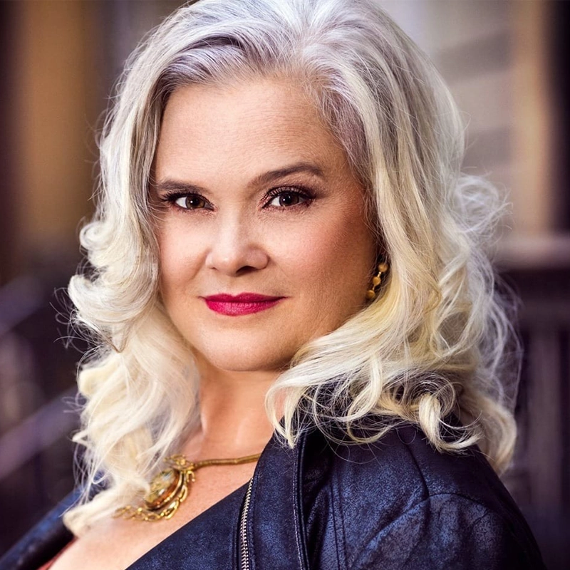 Julie Reyburn, with wavy, shoulder-length silver hair and fair skin, looks confidently at the camera. She wears bold red lipstick, gold earrings, a gold necklace, and a black leather jacket against a softly blurred brown and gray background.