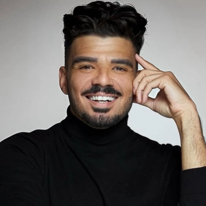 Julius Anthony Rubio, a man with dark hair and a trimmed beard, smiles warmly. He wears a black turtleneck and rests his right hand on the side of his face. The plain, softly lit background highlights his friendly expression.