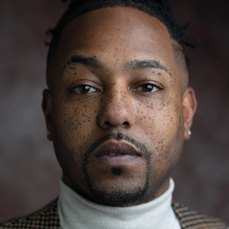 A close-up portrait of a man with short hair, facial hair, and many freckles—reminiscent of Dwayne Washington. He wears a light turtleneck sweater and a checkered jacket, looking directly at the camera with a neutral expression. The background is out of focus and dark.