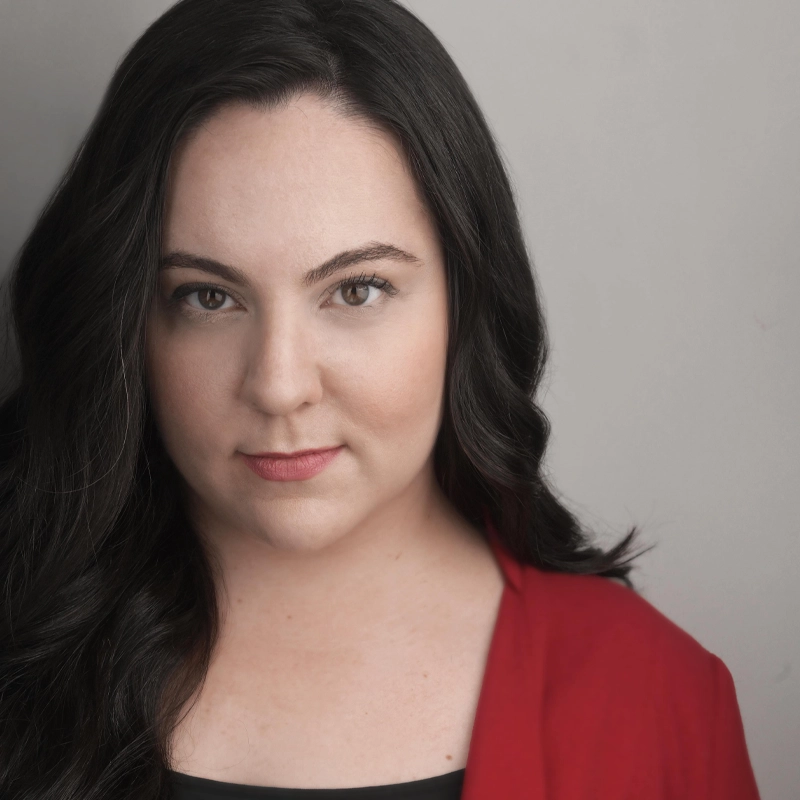 A woman with long, wavy dark hair and fair skin gazes confidently at the camera. Theresa Burns wears a red blazer over a black top, with subtle makeup and a neutral background, focusing on her face and upper shoulders.