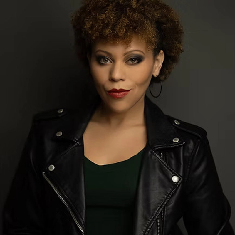 Kris Roberts, a woman with curly brown hair, wears hoop earrings, a dark green top, and a black leather jacket. She looks confidently at the camera with a slight smile, bold makeup, and stands against a dark, neutral background.