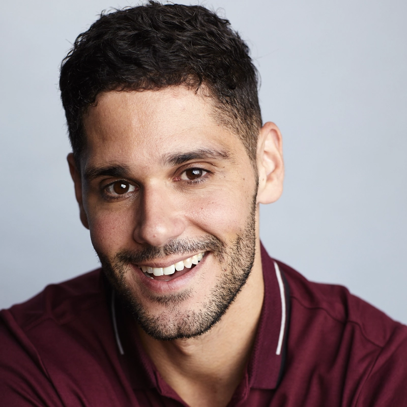 A smiling man, Perry Sherman, with short, dark curly hair and a trimmed beard wears a maroon collared shirt with white piping. He is facing the camera against a plain light gray background, creating a warm and friendly portrait.