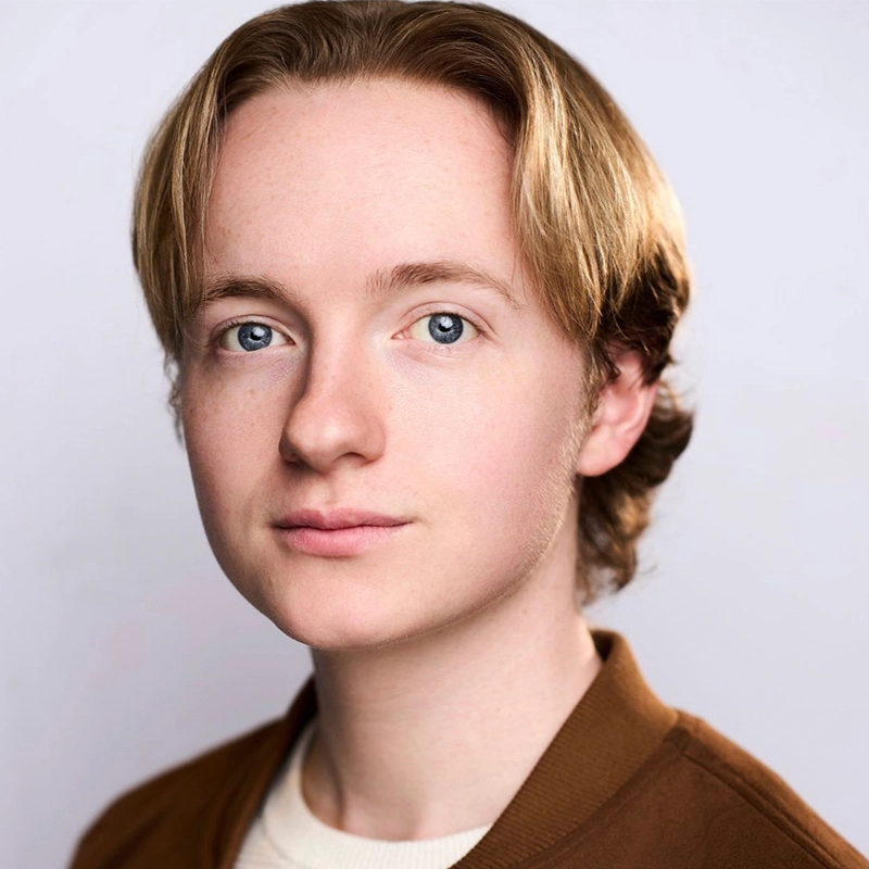 Madeline Smith is a young person with fair skin, blue eyes, and chin-length blond hair parted in the middle. Wearing a brown jacket over a white top, they face the camera with a neutral expression against a light gray background.