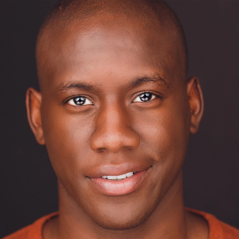 A close-up photo of a young person with dark skin, short hair, and brown eyes, smiling gently while looking directly at the camera. The background is plain and dark, making Sidney DuPont’s face the main focus.
