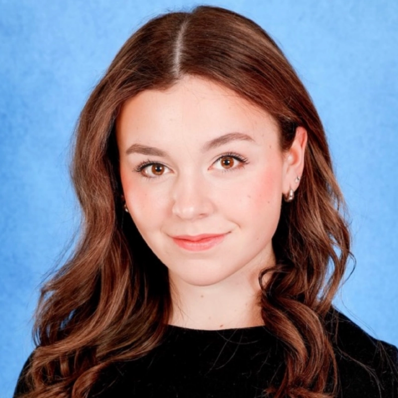 Madeline Smith, a young woman with long, wavy brown hair and fair skin, looks at the camera, smiling softly. She is wearing a black top, small earrings, and natural makeup against a solid bright blue background.