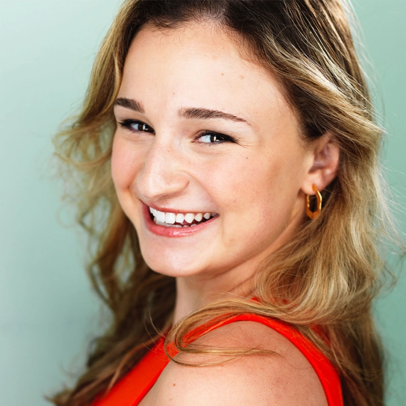 Madeline Smith, a young woman with wavy blonde hair, smiles brightly at the camera. She is wearing a sleeveless orange top and small gold hoop earrings. The soft light green background creates a vibrant and cheerful atmosphere.