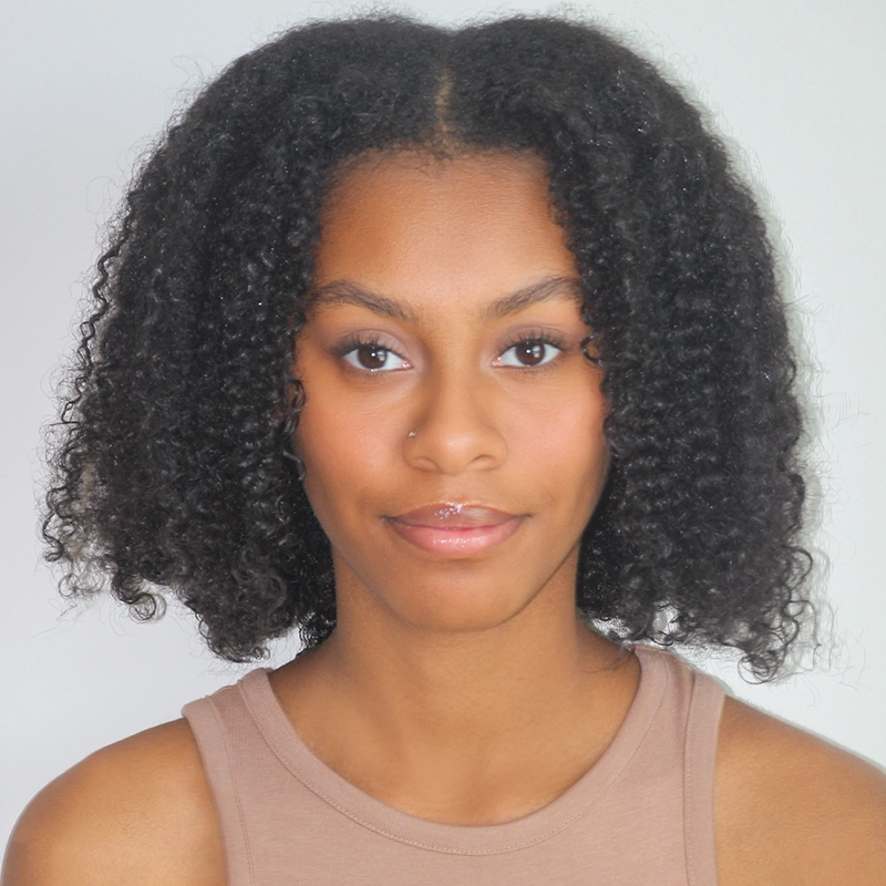 A young woman with medium-brown skin and natural, shoulder-length, curly black hair looks directly at the camera. Madeline Smith wears a sleeveless beige top and a nose stud, her neutral expression softened by a slight smile. The background is plain light gray.
