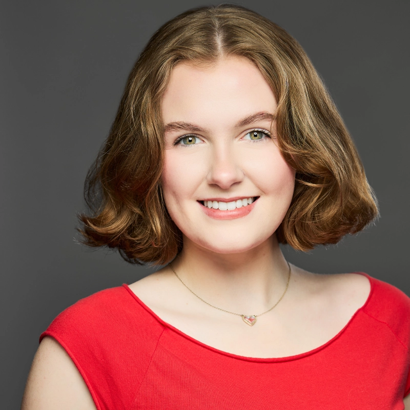 Grace Kennedy, a young woman with short, wavy, light brown hair, smiles at the camera. She wears a red top and a delicate necklace with a small heart pendant against a neutral gray background softly lit.