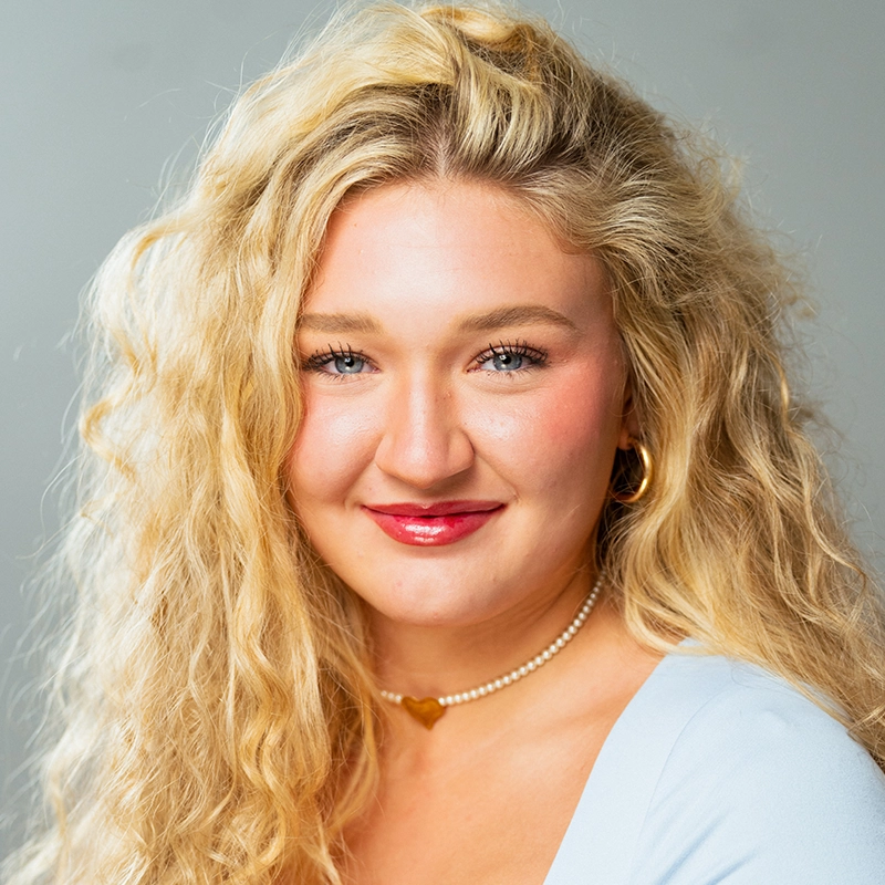 Madeline Smith, a young woman with long, wavy blonde hair, smiles gently. She has blue eyes, red lipstick, hoop earrings, a pearl necklace with a gold heart pendant, and wears a light blue top against a softly lit background.