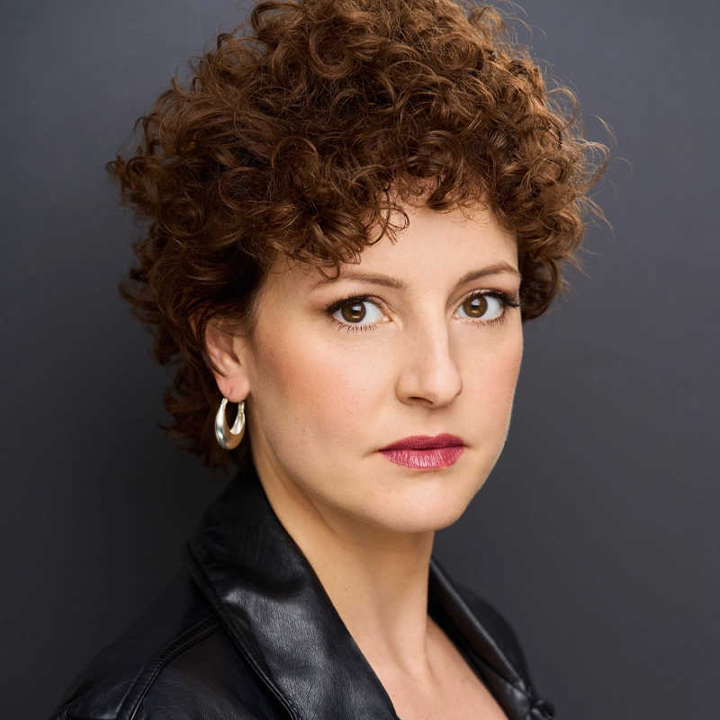 Andrea Prestinario, with short, curly brown hair and gold hoop earrings, stares directly at the camera. She has fair skin, neutral makeup with bold pink lipstick, and wears a black leather jacket against a dark gray background.