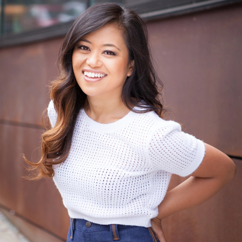 Renée Albulario, a young woman with long, dark wavy hair, smiles brightly at the camera. She wears a white short-sleeve textured knit top and high-waisted blue jeans, standing outdoors against a brown metal wall with her hands behind her back.