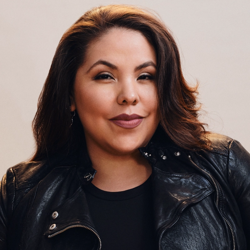 A woman with long, wavy brown hair smiles confidently. She is wearing a black leather jacket over a black top. The plain, light background and relaxed pose give this Scott Redmond portrait a modern yet approachable feel.