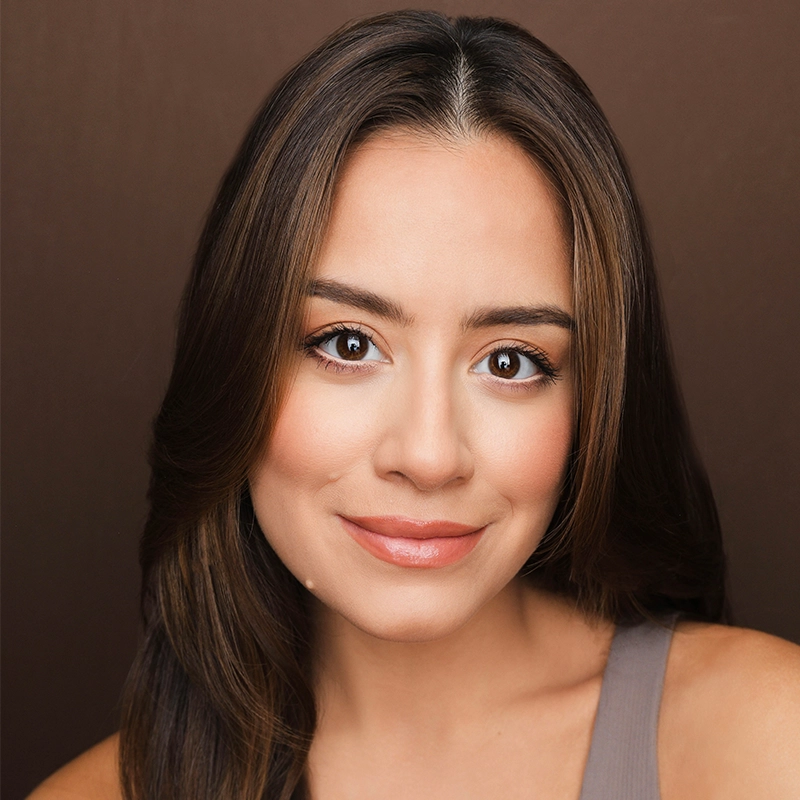 A woman with long, straight brown hair and brown eyes smiles gently at the camera. She wears natural makeup and a taupe sleeveless top. The Scott Redmond-inspired background is a plain, medium brown, creating a warm and soft portrait style.