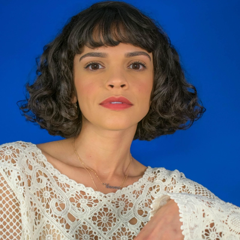 Gizel Jiménez, with short, curly dark hair and bangs, poses in front of a vibrant blue background. She wears a white crocheted top and a delicate gold necklace. Her expression is calm and confident, with natural makeup, pink blush, and lipstick.