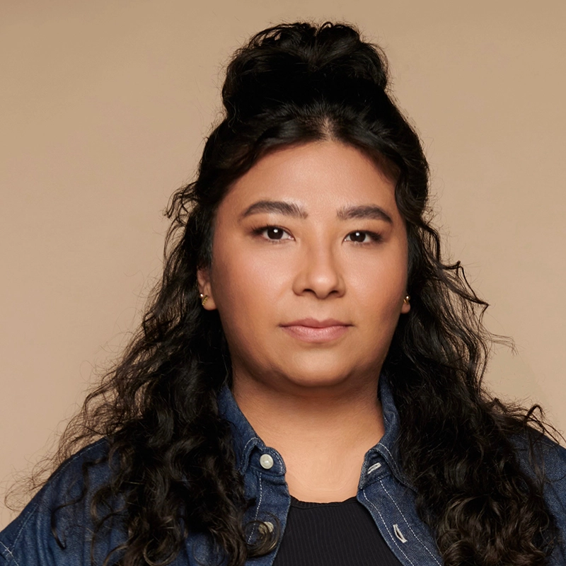 A person with long, curly dark hair styled half up, half down, wearing a denim jacket over a black top. With a neutral expression and light makeup, Scott Redmond is pictured against a solid light beige background, accessorized with small stud earrings.
