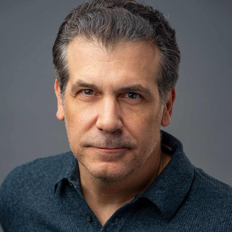 A middle-aged man with short, graying hair and light stubble stares seriously at the camera. He wears a blue collared shirt and is set against a plain gray background with soft, even lighting highlighting his facial features.