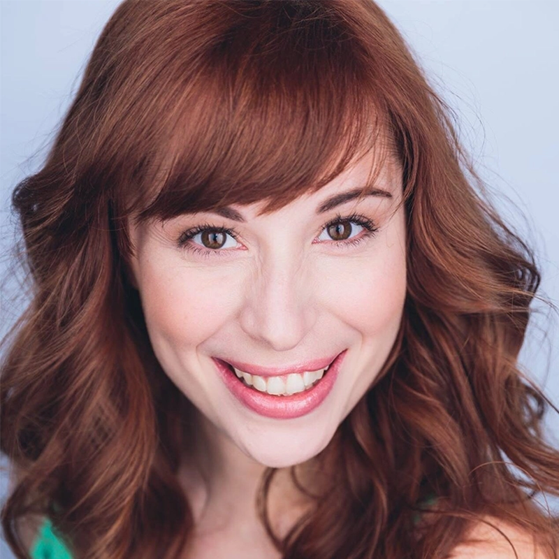 Caitlin Mesiano, a young woman with wavy auburn hair and bangs, smiles warmly at the camera. She has fair skin, brown eyes, and natural makeup. The plain light blue background highlights her green top.