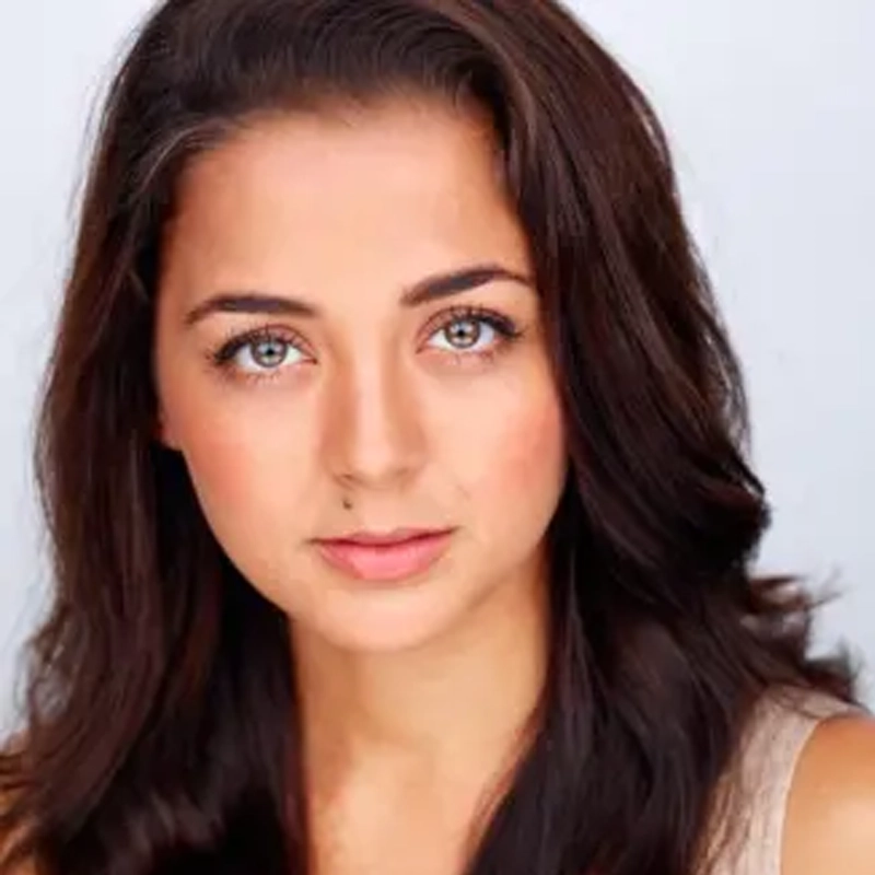 Claudillea Holloway, a woman with long, wavy dark brown hair and blue-green eyes, looks directly at the camera. She has light skin, natural makeup, and wears a light-colored sleeveless top against a softly lit background.