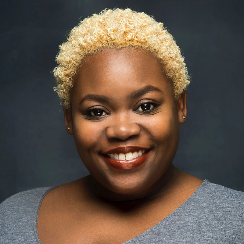A person with short, curly bleached blonde hair smiles warmly. They have brown skin, wear small earrings, and have a natural makeup look with red lipstick. Their top is gray and the background is a dark, solid color—capturing a style reminiscent of Sandra Okuboyejo’s vibrant confidence.