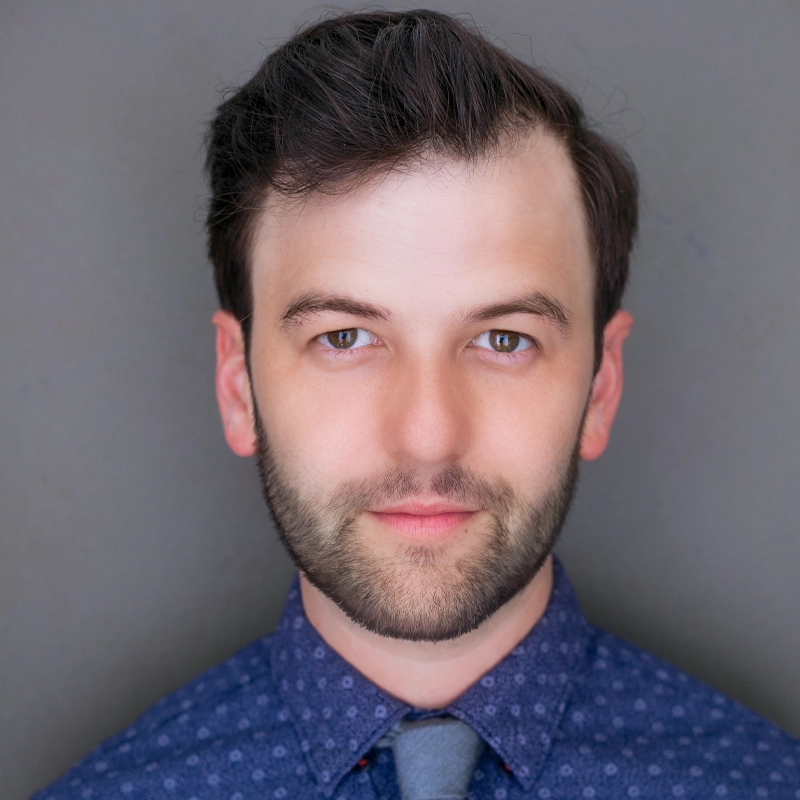 A man with fair skin, short dark hair, and a short beard looks directly at the camera with a neutral expression. He is wearing a blue patterned button-up shirt and a gray tie. The background is plain and gray.