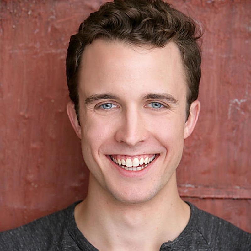 Eric Michael Parker, a young man with short, wavy brown hair and blue eyes, smiles broadly. He wears a dark grey t-shirt and stands against a textured reddish-brown wall. The image is well-lit, highlighting his cheerful expression.
