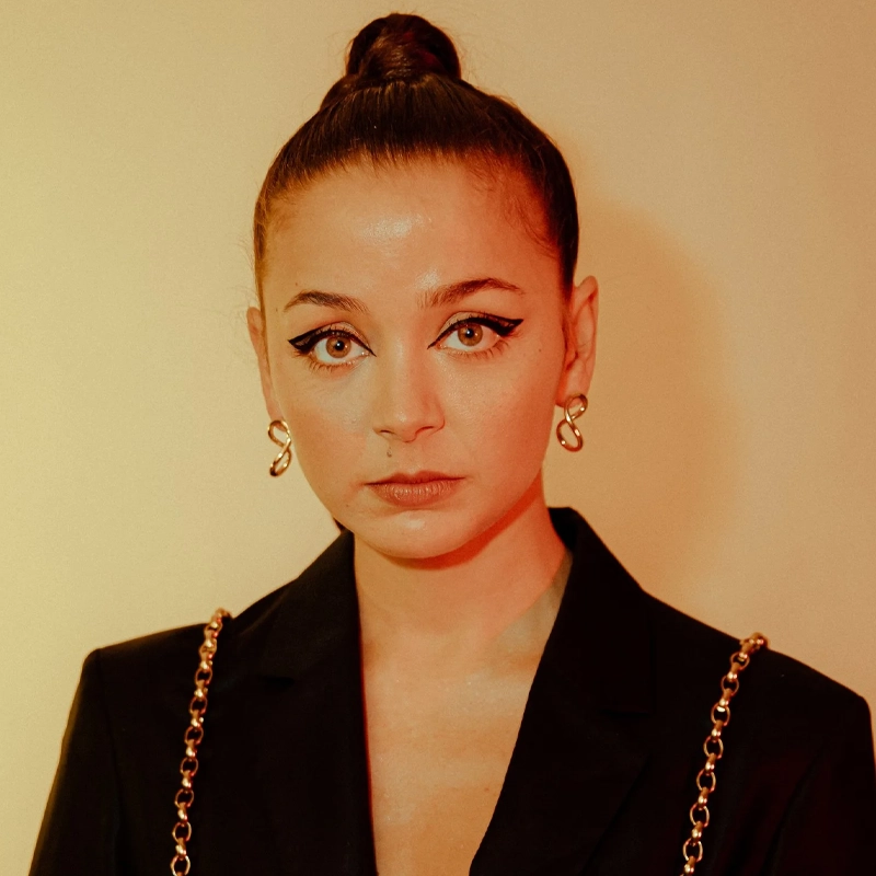 A woman with her brown hair in a high bun looks at the camera. Claudillea Holloway wears bold black winged eyeliner, gold hoop earrings, and a black blazer with gold chain straps. The background is a warm beige with soft reddish-yellow light.