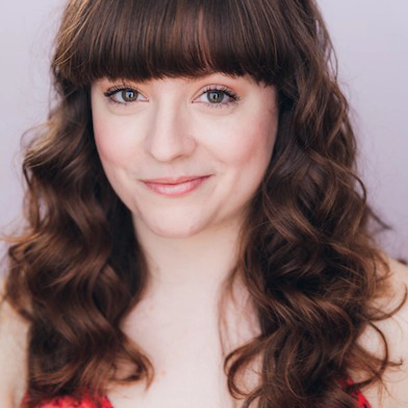 Cori Jaskier, a young woman with long, wavy brown hair and bangs, smiles softly at the camera. She has fair skin, blue eyes, and is wearing a red top against a plain light lavender background.
